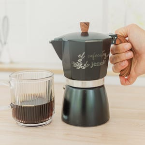 Puede incluir: Una cafetera italiana negra con asa y pomo de madera, junto a una taza de cristal transparente llena de café. La cafetera tiene el texto "el cafecito de Jennie". La taza tiene estrías verticales.