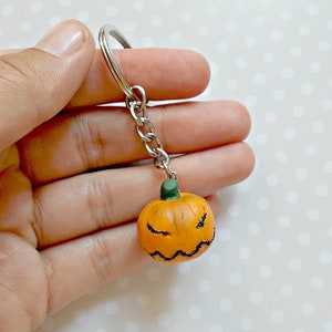 May include: A small orange pumpkin with a black jack-o'-lantern face and a green stem attached to a silver key chain.