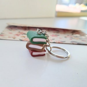 Peut inclure: Un porte-clés avec une pile de trois livres miniatures en pâte polymère. Les livres sont vert, marron et rouge avec des pages blanches. Le porte-clés est attaché à un anneau en métal argenté.