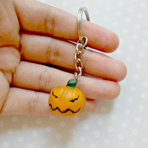 May include: A small orange pumpkin keychain with a green stem and a carved angry face. The keychain is attached to a silver metal chain and ring.