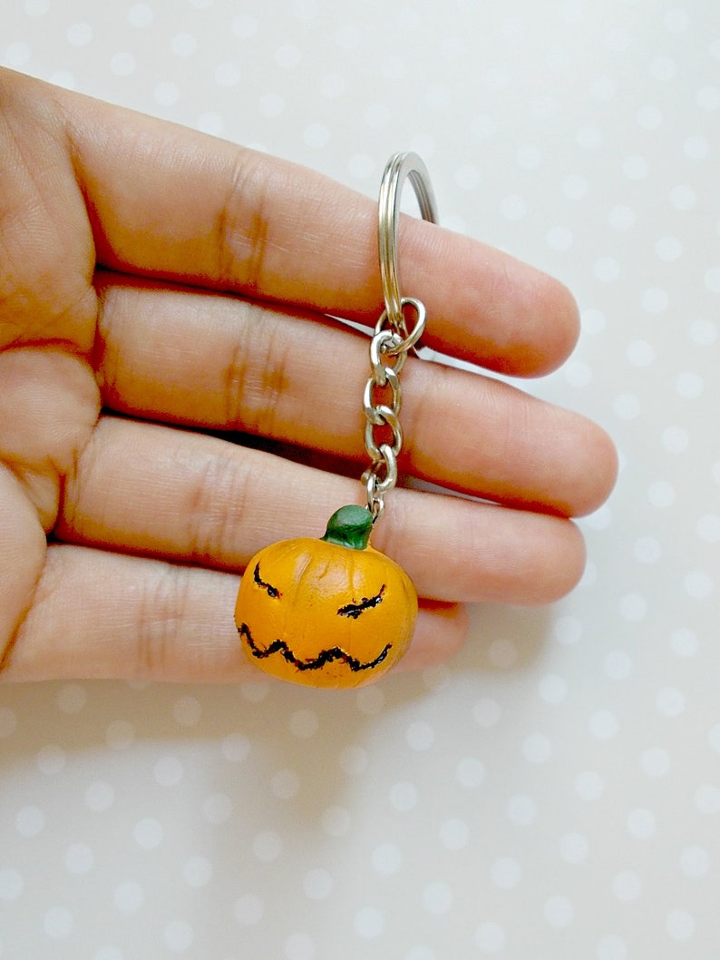 May include: A small orange pumpkin keychain with a green stem and a carved angry face. The keychain is attached to a silver metal chain and ring.