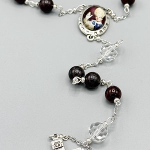 Puede incluir: Un rosario de plata con cuentas de madera de color rojo oscuro y un crucifijo de plata. La pieza central es un óvalo de plata con una imagen de un hombre con una túnica blanca. El rosario también tiene un óvalo de plata con una imagen de la Virgen María.