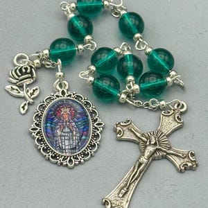 May include: A rosary with green glass beads and silver-toned metal accents. The rosary includes a decorative pendant with an image of a religious figure, a silver cross, and a rose charm.