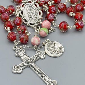 May include: A rosary with red and pink floral beads, silver-toned crucifix, and a pendant with a rose design. The beads have a glass-like appearance. The rosary also features a silver-toned centerpiece with a religious image.