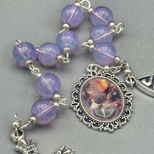 May include: A rosary with lavender-colored beads and silver-toned metal accents. The rosary features an oval pendant with a religious image and a detailed crucifix.
