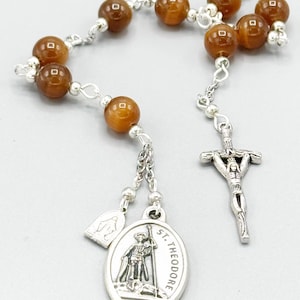 Pode incluir: Um rosário de contas de olho de tigre castanho com um crucifixo de prata e um pingente oval de prata com um santo. O pingente diz "St. Theodore".
