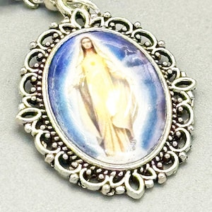 May include: Silver-toned oval pendant featuring a religious image of a woman in a flowing gown, set against a blue and yellow background. The pendant has an ornate, filigree-style border and is suspended from a chain.