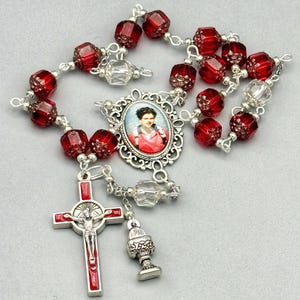 May include: A rosary with red faceted beads and silver-toned metal accents. The rosary features a crucifix, a small chalice charm, and a central oval pendant with a portrait. The beads are approximately 0.5 inches in diameter.