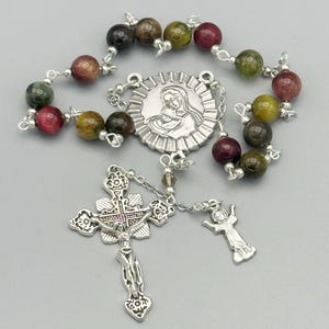 Puede incluir: Un rosario con cuentas de piedras preciosas multicolores, un crucifijo de plata y un colgante de plata con la imagen de una figura religiosa. Las cuentas son una mezcla de verde, marrón y rojo. El rosario está sobre una superficie gris.