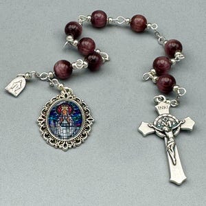 Peut inclure: Un chapelet avec des perles rouge foncé et des accents métalliques argentés. Le chapelet comprend un pendentif ovale décoratif avec une image religieuse et un crucifix avec l'inscription "INRI".
