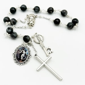 May include: A black rosary with silver beads and charms. The charms include a cross, a key, and a medal with a picture of the Virgin Mary. The rosary is on a white background.