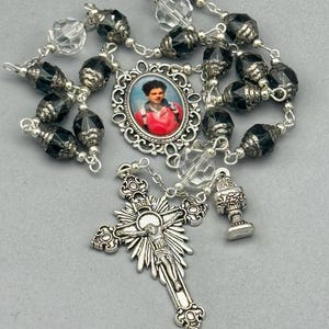 May include: A rosary with black faceted beads, clear crystal beads, and silver-toned metal accents. The centerpiece features an oval portrait, and the cross depicts Jesus. A chalice charm is also present.