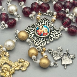 May include: A rosary featuring translucent deep red beads, white pearl-like beads, and matte gold-toned beads. It includes a silver-toned centerpiece with a circular image of Jesus and Mary, a gold-toned crucifix with INRI, and two heart charms.