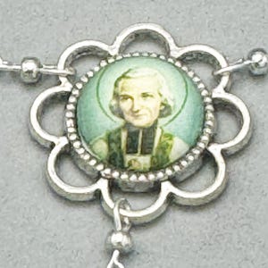 May include: A silver-toned religious pendant featuring a portrait of a saint within a flower-shaped frame. The portrait is set against a light green background. The pendant is connected to a chain with small, round beads.