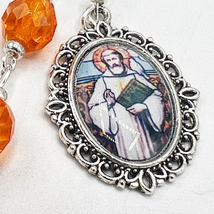 May include: A silver rosary chain with orange glass beads and a silver oval pendant featuring a stained glass image of a saint holding a book.