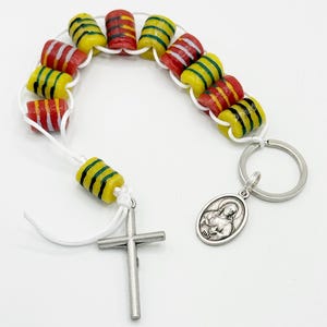 May include: A white cord with a silver cross charm and a silver key chain with a silver oval medal featuring a religious image. The cord has a series of yellow, red, and green glass beads.