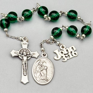 Peut inclure: Un chapelet composé de perles en verre vert émeraude translucide reliées par une chaîne en métal argenté. Il comprend un crucifix argenté avec l'inscription INRI, une médaille ovale représentant une scène religieuse et un pendentif John 3:16.