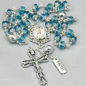 May include: A rosary with blue faceted beads and silver-toned accents. The rosary features a silver crucifix with a figure of Jesus and a pendant that reads "FAITH". A central medallion depicts a religious image.