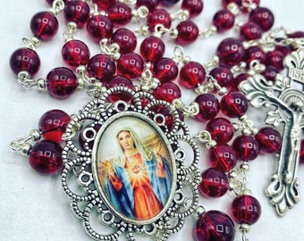 May include: A rosary with deep red glass beads and silver-toned metal accents. The centerpiece features an oval image of a religious figure within an ornate frame. A silver crucifix is also present.
