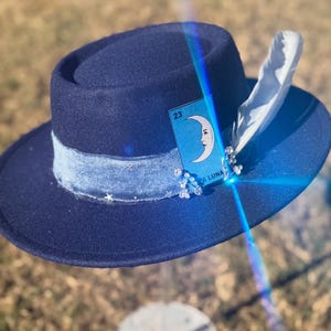 May include: A navy blue fedora hat with a light blue velvet band and a white feather. The hat is decorated with a card featuring a crescent moon and the number 23, along with small star and bead accents.
