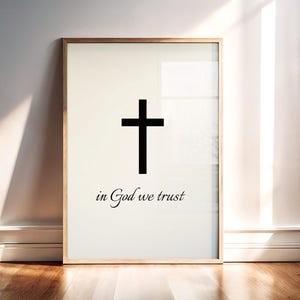 Może przedstawiać: Oprawiony wydruk z czarnym krzyżem na kremowym tle. Pod krzyżem napisano "in God we trust" czcionką skryptową. Rama jest w kolorze jasnego drewna, a grafika jest na białej ścianie.