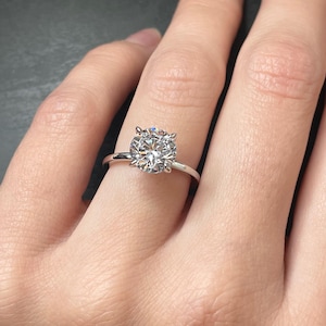 May include: A solitaire engagement ring with a large, round, clear gemstone set in a thin, silver band. The ring is displayed on a person's hand, showcasing the sparkling stone. The ring's design is simple and elegant.
