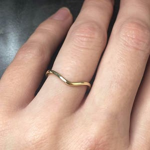 May include: A gold-colored ring with a curved, wave-like design. The ring is smooth and shiny, reflecting light. The ring is worn on a finger, showcasing its elegant and minimalist style. This piece of jewelry is suitable for everyday wear.
