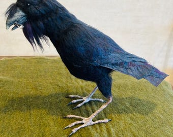 May include: A handcrafted, dark blue raven sculpture with a textured, feathered appearance. The bird has a detailed beak, piercing eyes, and stands on detailed feet. The raven's feathers have hints of purple and blue.