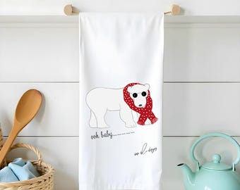 May include: A white kitchen towel featuring a graphic illustration of a polar bear wearing black sunglasses and a red scarf with white polka dots. The text "ooh baby............" is printed in black below the bear, along with a signature script.