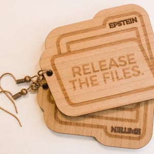 May include: Wooden earrings in the shape of file folders. The earrings are light brown with the text "RELEASE THE FILES" and "EPSTEIN" engraved on them. The earrings have copper-colored ear wires.