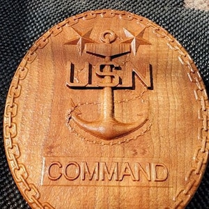 Può includere: Targa rotonda in legno con un design intagliato. Il design include le lettere "USN" sopra un'ancora, due stelle e la parola "COMMAND". La targa è bordata da un motivo a catena.