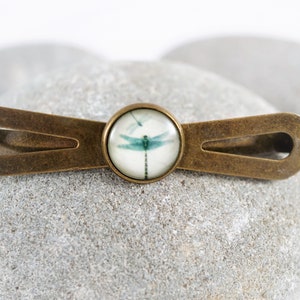 May include: A bronze tie clip with a glass cabochon featuring a blue dragonfly on a white background.