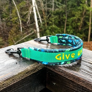 Puede incluir: Una correa para perros verde y azul con cierres metálicos negros. La correa tiene un patrón de dinosaurios y las palabras "Give" en amarillo. La correa está sobre una superficie de madera.