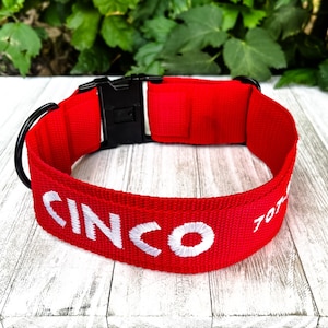 May include: A bright red nylon pet collar featuring the embroidered white text CINCO and partial numbers on the side. The collar includes a black plastic side-release buckle and a black metal D-ring for attachment, presented in a circular shape.