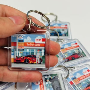 Op de afbeelding: Een transparante sleutelhanger van plastic met een rode auto voor een Tim Hortons en een Esso-tankstation. De sleutelhanger heeft een cartoonillustratie van een persoon die naast de auto staat. De tekst "Esso Tim Hortons" staat op de sleutelhanger gedrukt.