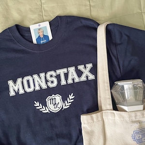 May include: A navy blue t-shirt with the word "MONSTAX" printed in white on the front. The shirt also features a white logo with a stylised "M" and a sword. The shirt is laid out on a beige tote bag with a white logo.