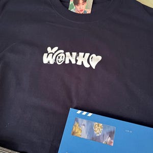 May include: A navy blue t-shirt with the word "WONKO" in white, incorporating a smiley face and heart. A photo card with a person wearing a pink bow is on the shirt. A blue book with "Blue Letter" is also visible.