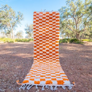 Könnte beinhalten: Ein weiß-orangener Schachbrettteppich mit Fransenbordüre. Der Teppich ist aus weichem, flauschigem Material gefertigt und perfekt, um jedem Raum einen Hauch von Farbe und Textur zu verleihen.