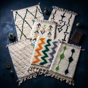 Op de afbeelding: Een collectie van zes rechthoekige kleden van crèmekleurige wol met diverse patronen, waaronder donkergroene ruiten, zwarte geometrische pijlen, veelkleurige zigzaglijnen, diagonale ribbels en verticale lijnen, allemaal afgewerkt met franjes.