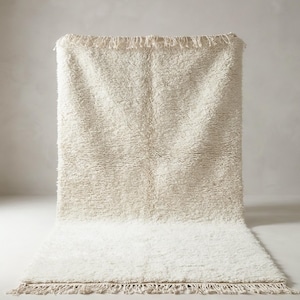 May include: A cream-colored, rectangular rug with a thick, textured pile. The rug features a fringe along the edges and a lighter shade of cream at the bottom. The rug is displayed against a neutral background.