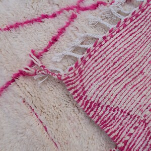 May include: A white and pink Moroccan rug with a geometric pattern. The rug has a fringe border and is made of wool.