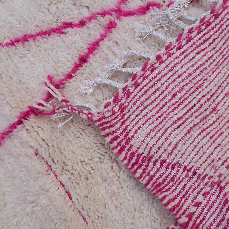 May include: A white and pink Moroccan rug with a geometric pattern. The rug has a fringe border and is made of wool.
