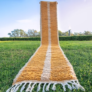 May include: A long, narrow, mustard yellow and white striped rug with fringe on the ends. The rug is laid out on a grassy lawn.
