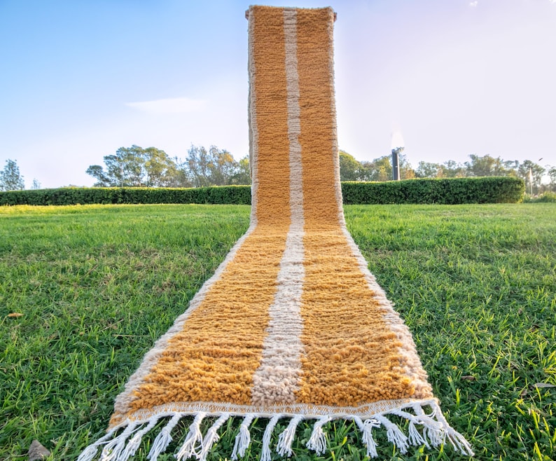 May include: A long, narrow, mustard yellow and white striped rug with fringe on the ends. The rug is laid out on a grassy lawn.