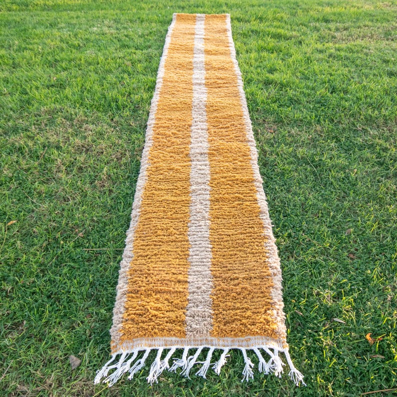 May include: A long, narrow, mustard yellow rug with two white stripes running down the centre. The rug has a fringe on both ends.