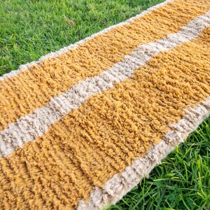 May include: A long, rectangular rug with a striped pattern in shades of beige and mustard yellow. The rug is made of a soft, fluffy material and has a slightly textured surface.