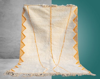 May include: A white Moroccan rug with a geometric pattern in orange. The rug has a fringe border and is made of wool.
