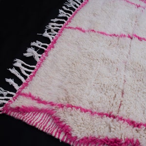 May include: White and pink Moroccan rug with a fringe border. The rug has a fluffy texture and a geometric pattern.
