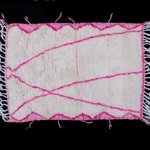 May include: White rug with a pink geometric design. The rug has a fringe border.