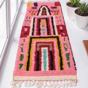 May include: Pink rectangular rug with a geometric design. The rug features a central rectangular pattern with black, yellow, red, and blue accents. The rug has a fringe on the bottom edge and is made of a textured material.